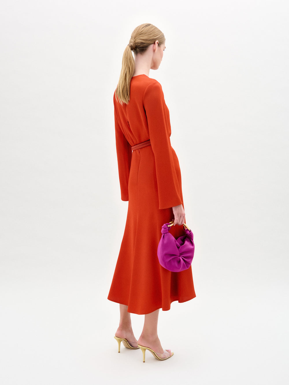 A person with light brown hair in a ponytail is wearing an ankle-length A-line orange Pesaro Dress by SVT DESIGNS and strappy heels, holding a bright purple handbag against a plain light background.