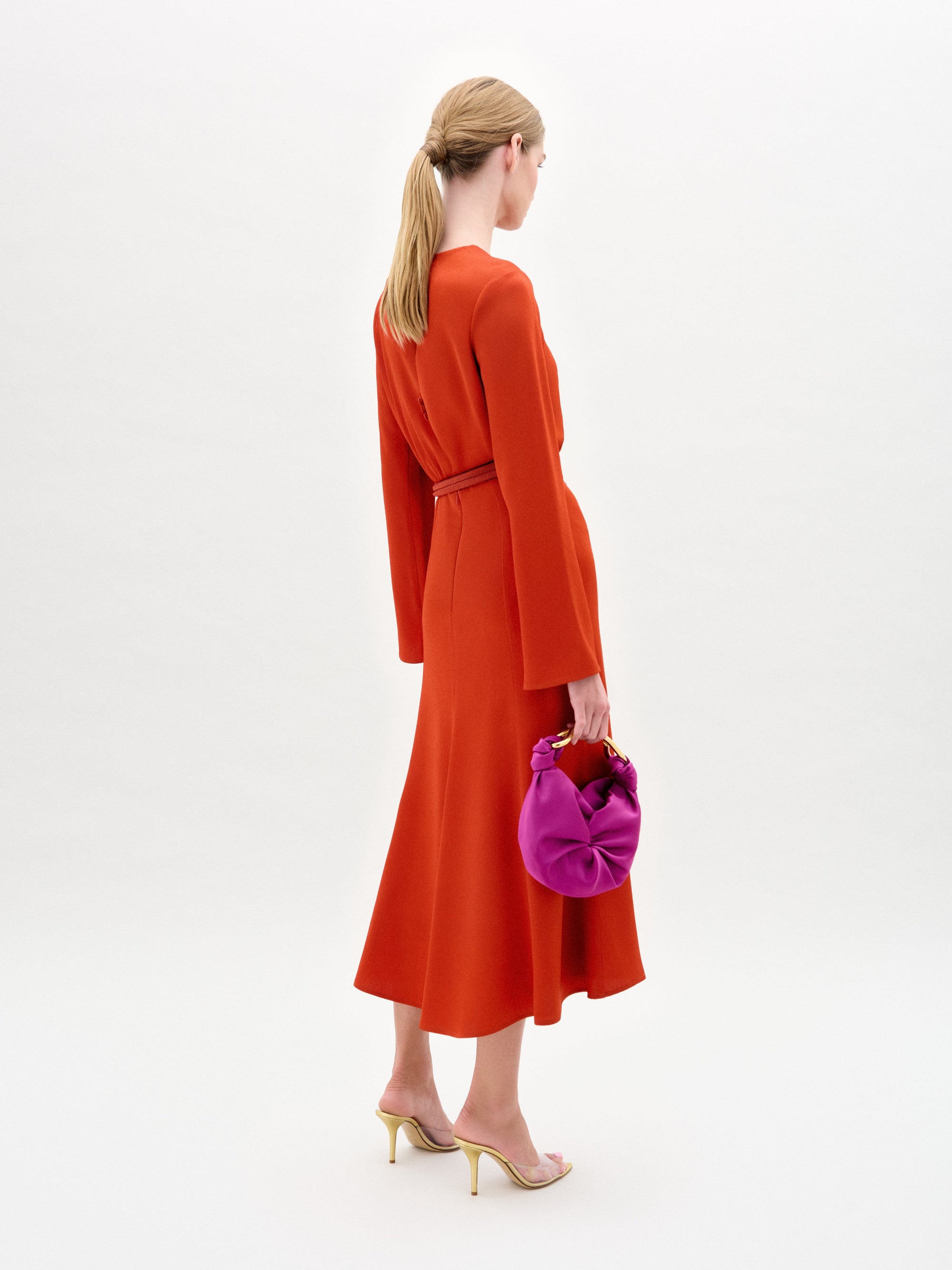 Woman wearing the Pesaro Dress, a long-sleeved orange midi dress with a belted waist crafted from premium Italian fabric, paired with a small purple bag and transparent high-heeled sandals, against a plain white background.