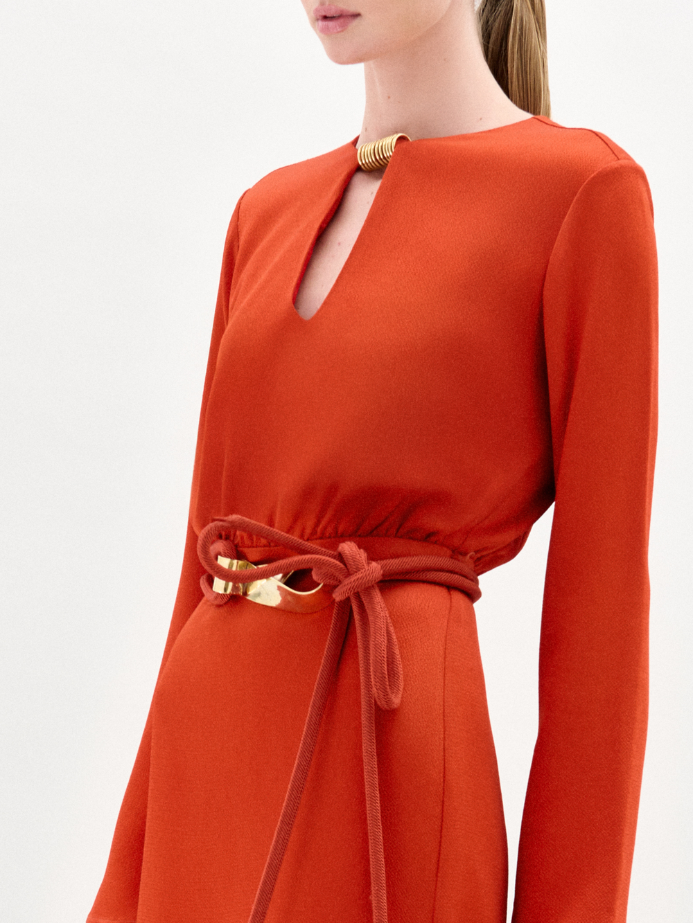 Woman wearing the Pesaro Dress, a long-sleeved orange midi dress with a belted waist crafted from premium Italian fabric, paired with a small purple bag and transparent high-heeled sandals, against a plain white background.