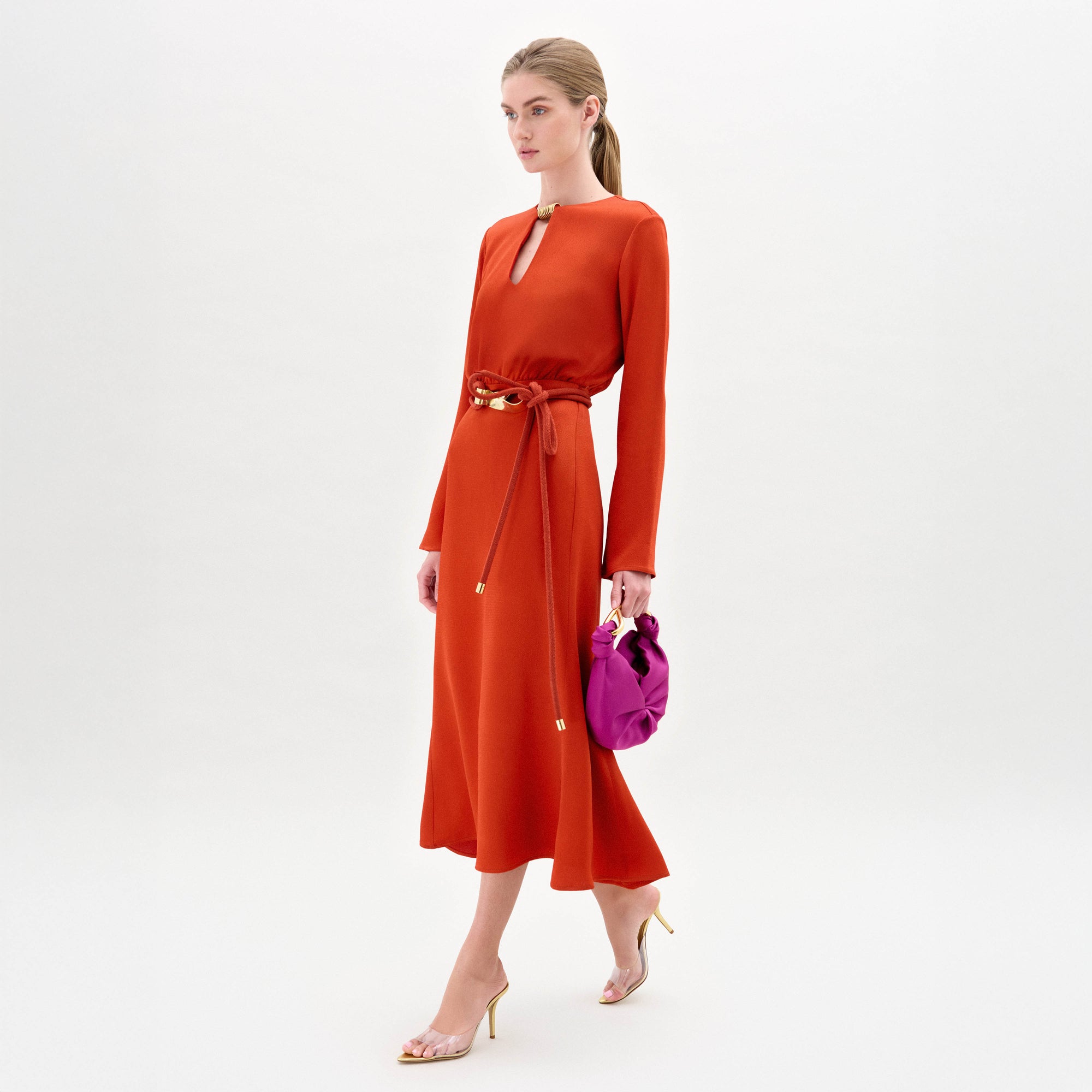 A woman poses against a white background in the Pesaro Dress by SVT DESIGNS, a long-sleeved orange midi dress of fine Italian fabric with a keyhole neckline and knotted belt. She pairs it with a purple handbag and clear heeled sandals.