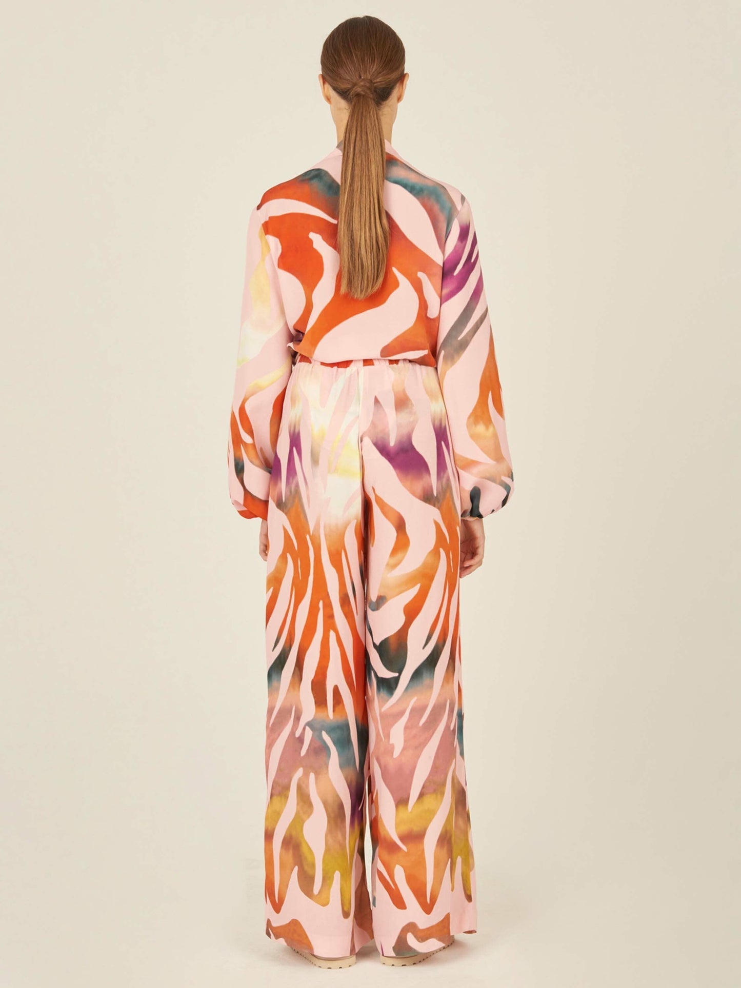 The Honey Blouse Blush Prismatic is a long-sleeve top with a vibrant abstract zebra print in pink, orange, yellow, and white. It features a front tie knot design and a flowy silhouette.