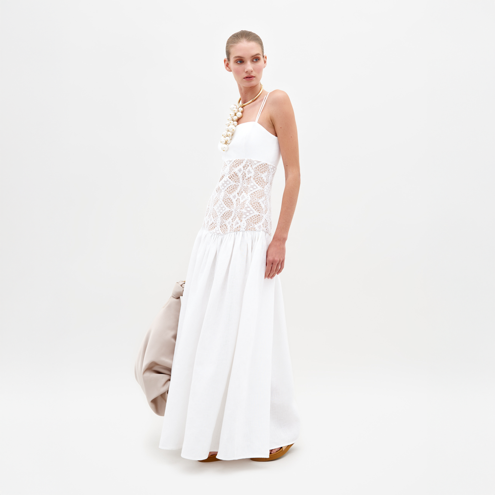 A woman stands against a plain white background, wearing the Pilar Dress—a flowing maxi with a lace bodice and guipure panel—paired with a large beige bag, slicked-back hair, and a chunky pearl necklace.