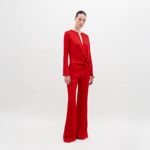 Against a plain white background, an individual models the SVT DESIGNS Palermo Pant. The red ensemble includes long sleeves and high-waisted, wide-leg pants with the top featuring a central knot detail, while they look directly at the camera.