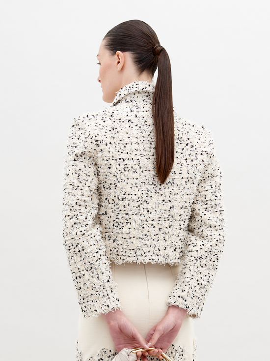 A woman with light skin and dark hair is wearing a Perla Jacket Beige Black Embroidered Flowers over a matching dress. Holding a small bag, she stands against a plain background. Pre-order now to ensure it ships by November 15th, 2024.