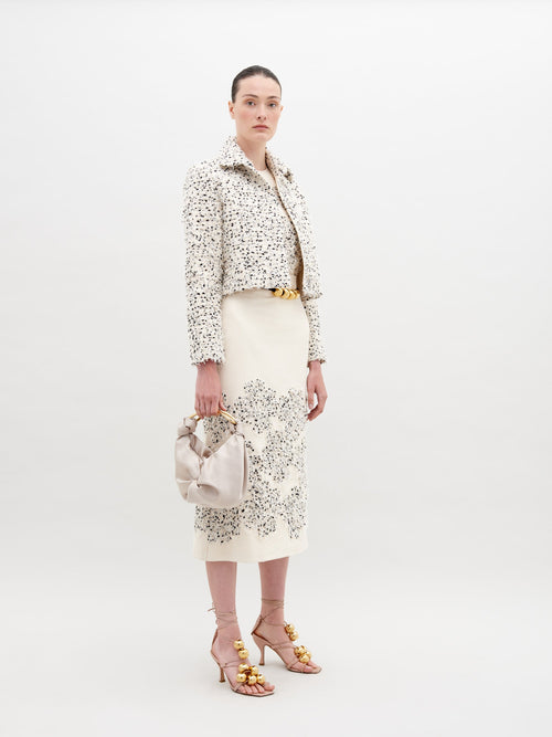 A woman stands wearing a white patterned jacket, Atira Skirt Beige Black Embroidered Flowers, gold belt, and gold-embellished sandals. She holds a beige handbag against a plain background. Don't miss out—pre-order now for the November 15th, 2024 ship-by date!