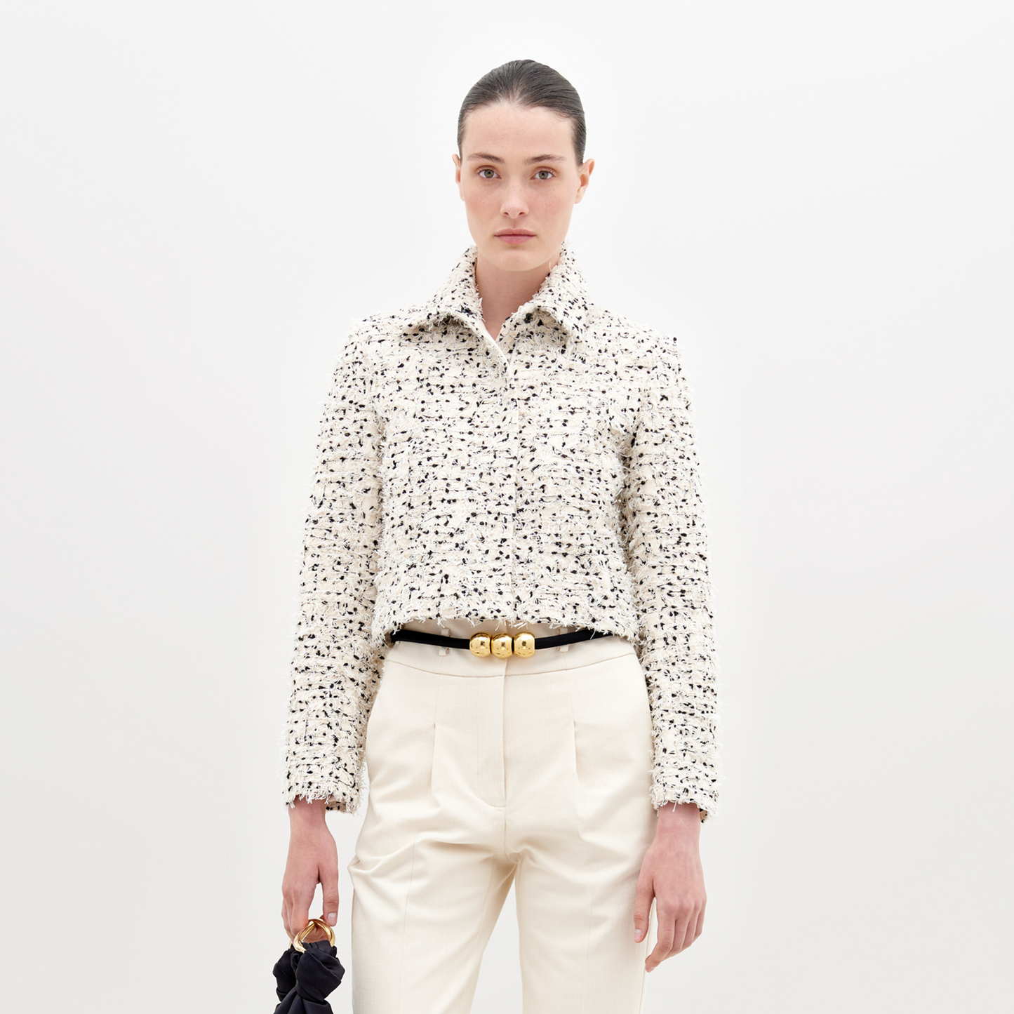 A woman poses against a white backdrop wearing the SVT DESIGNS Perla Jacket—a textured, long-sleeve cream blouse with black accents crafted from fine Italian textile—paired with cream high-waisted trousers, a black belt with gold accents, and holding a black item.