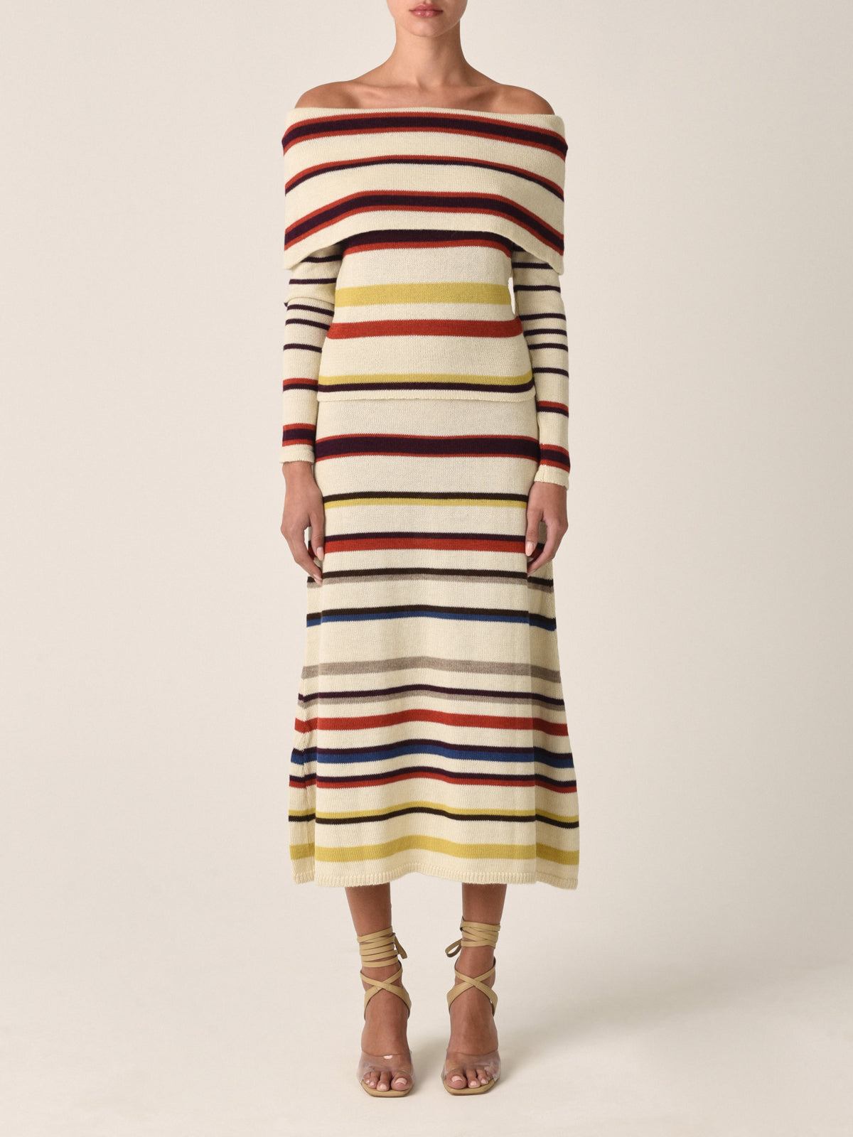 A person is elegantly styled in the Alexandra Knit Top Multi Mustard Stripe by Silvia Tcherassi, showcasing off-the-shoulder, multi-colored stripes including red, yellow, and blue. Made from 100% baby alpaca with a mid-calf drape, it's paired with strappy high-heeled sandals against a plain backdrop.