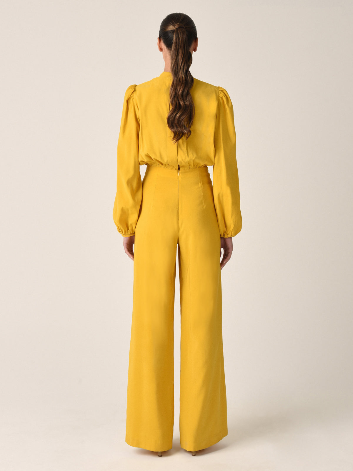 The Arnetia Jumpsuit Amber in mustard yellow features long puffed sleeves, a keyhole neckline, and a twisted knot detail at the front, capturing the versatile elegant appeal of the Silvia Tcherassi aesthetic.