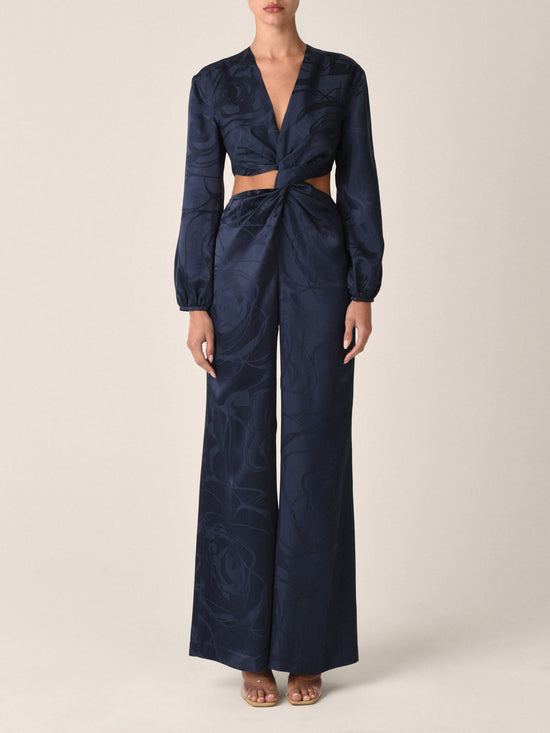 The Joely Jumpsuit Navy Jacquard, a black long-sleeve piece from the Resort 2023 collection, features a twisted front design, cutout at the waist, and wide-leg trousers.