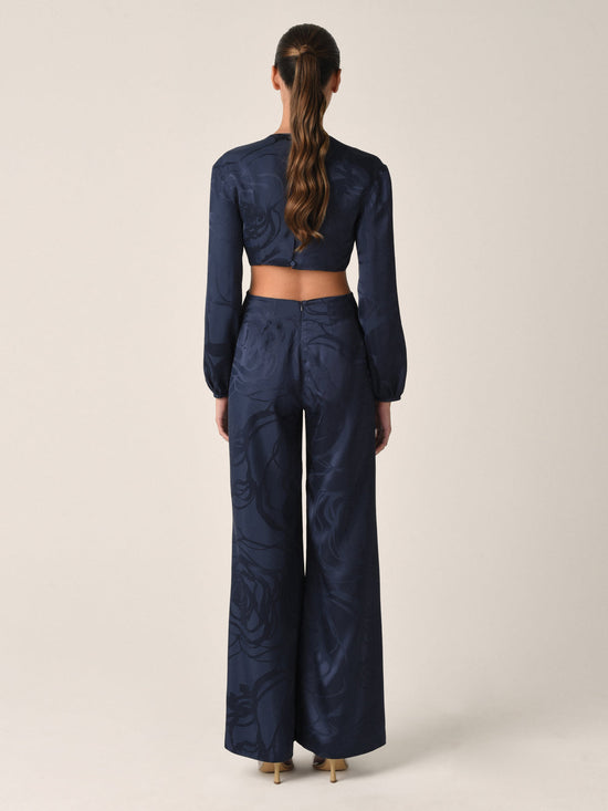 The Joely Jumpsuit Navy Jacquard, a black long-sleeve piece from the Resort 2023 collection, features a twisted front design, cutout at the waist, and wide-leg trousers.