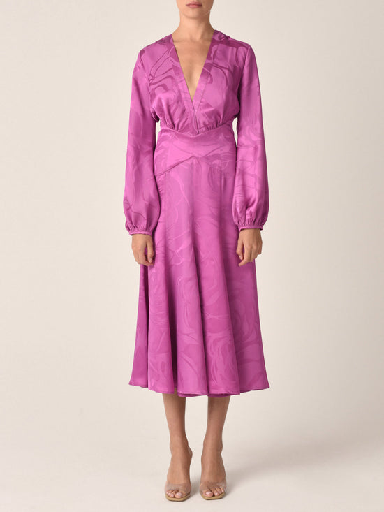 A long-sleeved, floor-length Kathryn Dress Magenta Orchid Jacquard with a V-neck and criss-cross bodice, featuring a subtle jacquard pattern and a tag near the neckline.