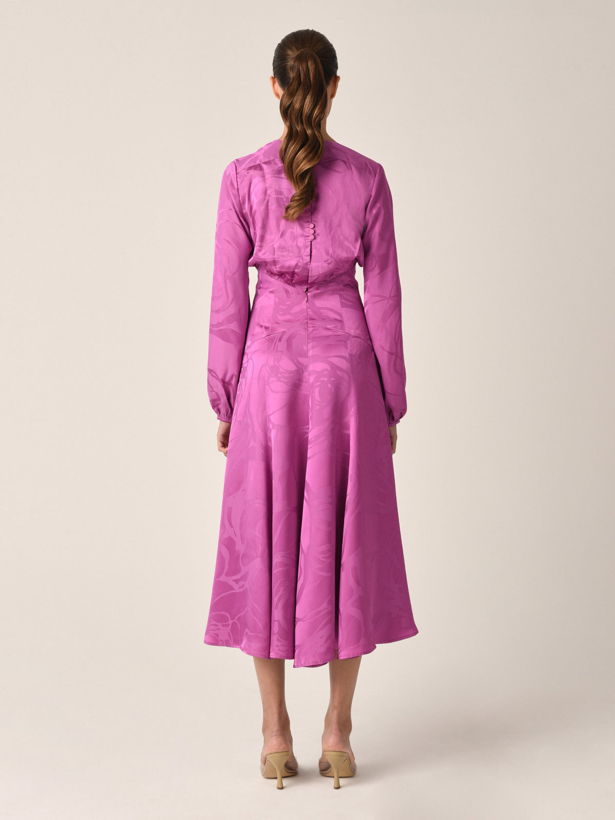 A long-sleeved, floor-length Kathryn Dress Magenta Orchid Jacquard with a V-neck and criss-cross bodice, featuring a subtle jacquard pattern and a tag near the neckline.