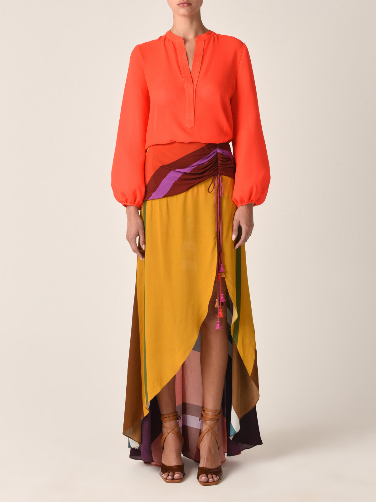 A person wears the Silvia Tcherassi Triora Blouse in tangerine, paired with a vibrant layered wrap skirt featuring yellow, brown, and purple hues. Strappy brown sandals complete the chic ensemble against a plain light backdrop.