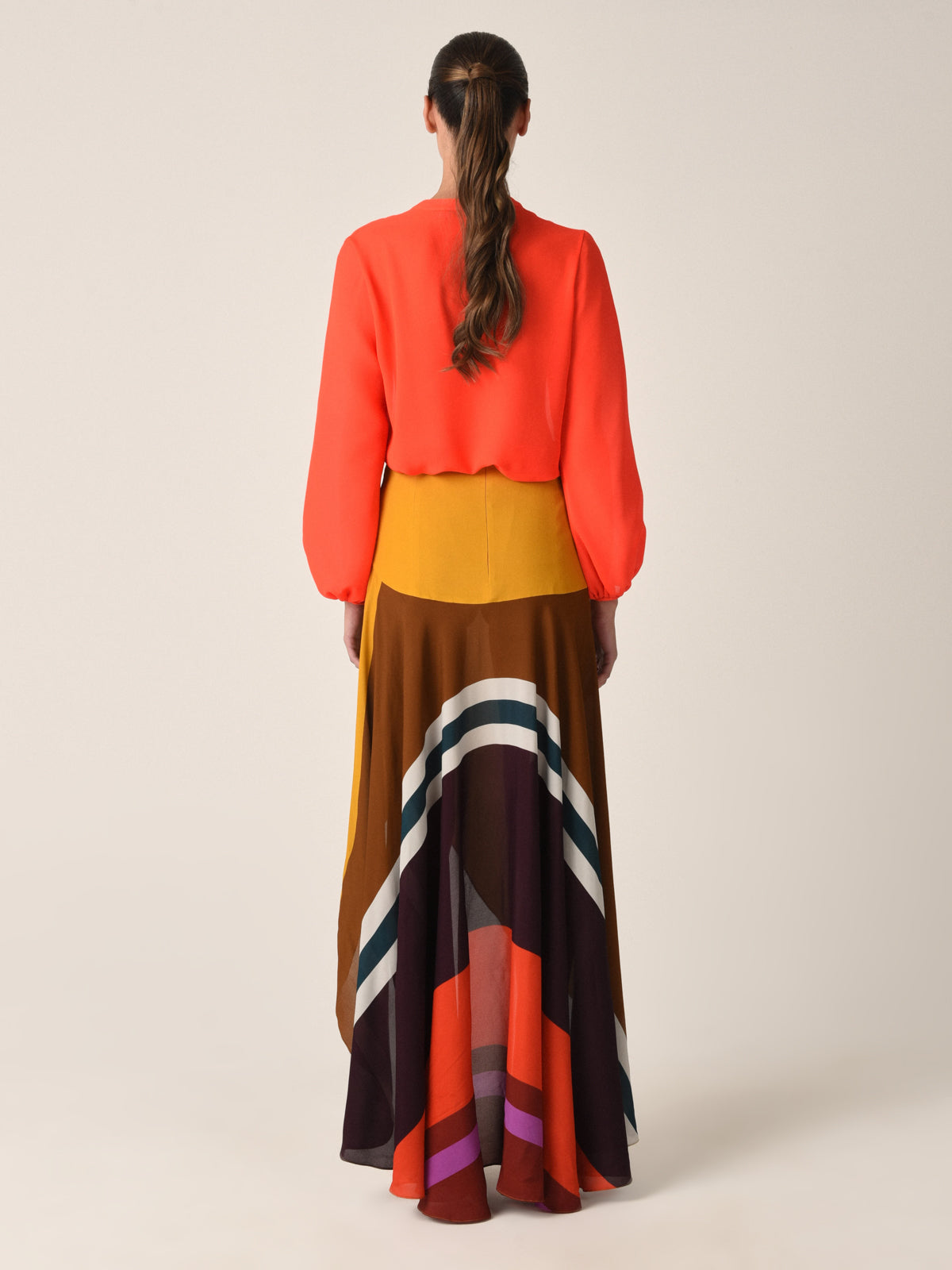 A woman with long hair in a low ponytail stands with her back to the camera, wearing the chic Triora Blouse in tangerine viscose by Silvia Tcherassi, featuring billowing sleeves. Her multicolored, asymmetrically layered skirt adds flair against the plain, light-colored background.