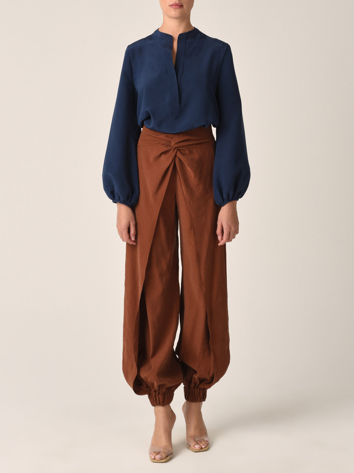 A person models the chic Silvia Tcherassi Triora Blouse in navy with billowing sleeves and pairs it seamlessly with high-waisted brown pants featuring gathered cuffs. They stand against a neutral background, wearing clear, open-toed shoes, while their face is obscured.