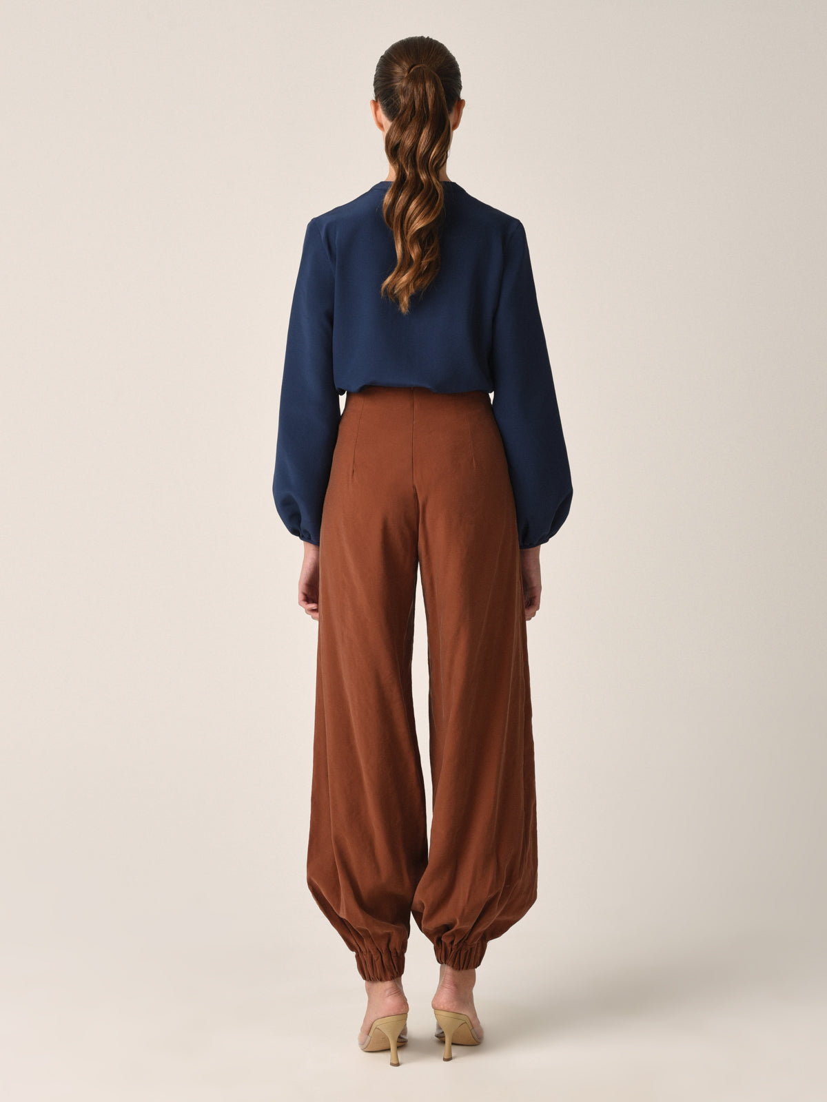 A woman faces away from the camera, wearing a Silvia Tcherassi Triora Blouse in navy with high-waisted brown tapered pants and beige heels. Her hair is styled in a ponytail, set against a plain light background that highlights her chic look.