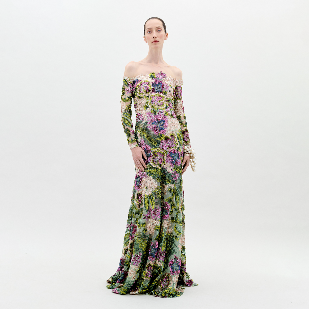 A person stands against a plain background wearing the Rafaella Dress Botanical Sequins, a floor-length, off-the-shoulder floral gown with long sleeves and adorned with a vibrant pattern of flowers and greenery. This elegant piece is available for pre-order now, with a ship-by date set for February 2025.