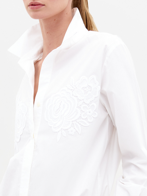 Sporting the Raizel Blouse White, a person with long hair stands against a plain background with hands in pockets, dressed in a white embroidered button-up shirt with the collar turned up and coordinating white pants. They are ready for the pre-order shoot scheduled for February 15th, 2025.