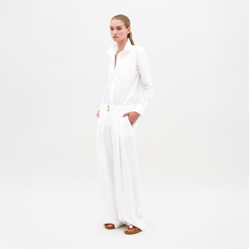 Against a plain backdrop, a person exudes effortless style wearing the SVT DESIGNS Opal Pant—a pair of high-waisted wide-leg trousers—and a white long-sleeve shirt with an embroidered design. With hands in pockets and sandals on their feet, they gaze thoughtfully to the side, showcasing Italian textile flair.