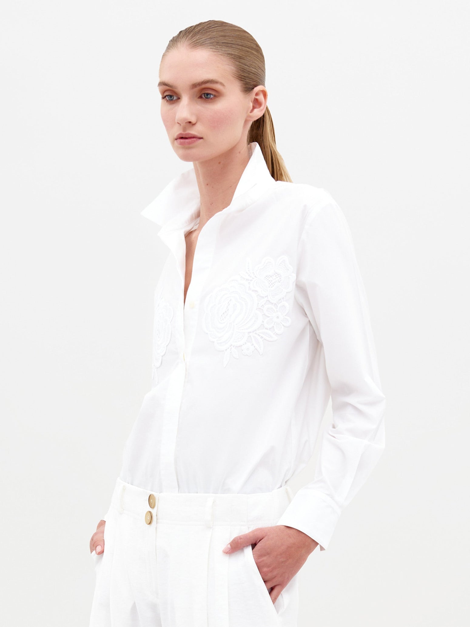 Sporting the Raizel Blouse White, a person with long hair stands against a plain background with hands in pockets, dressed in a white embroidered button-up shirt with the collar turned up and coordinating white pants. They are ready for the pre-order shoot scheduled for February 15th, 2025.