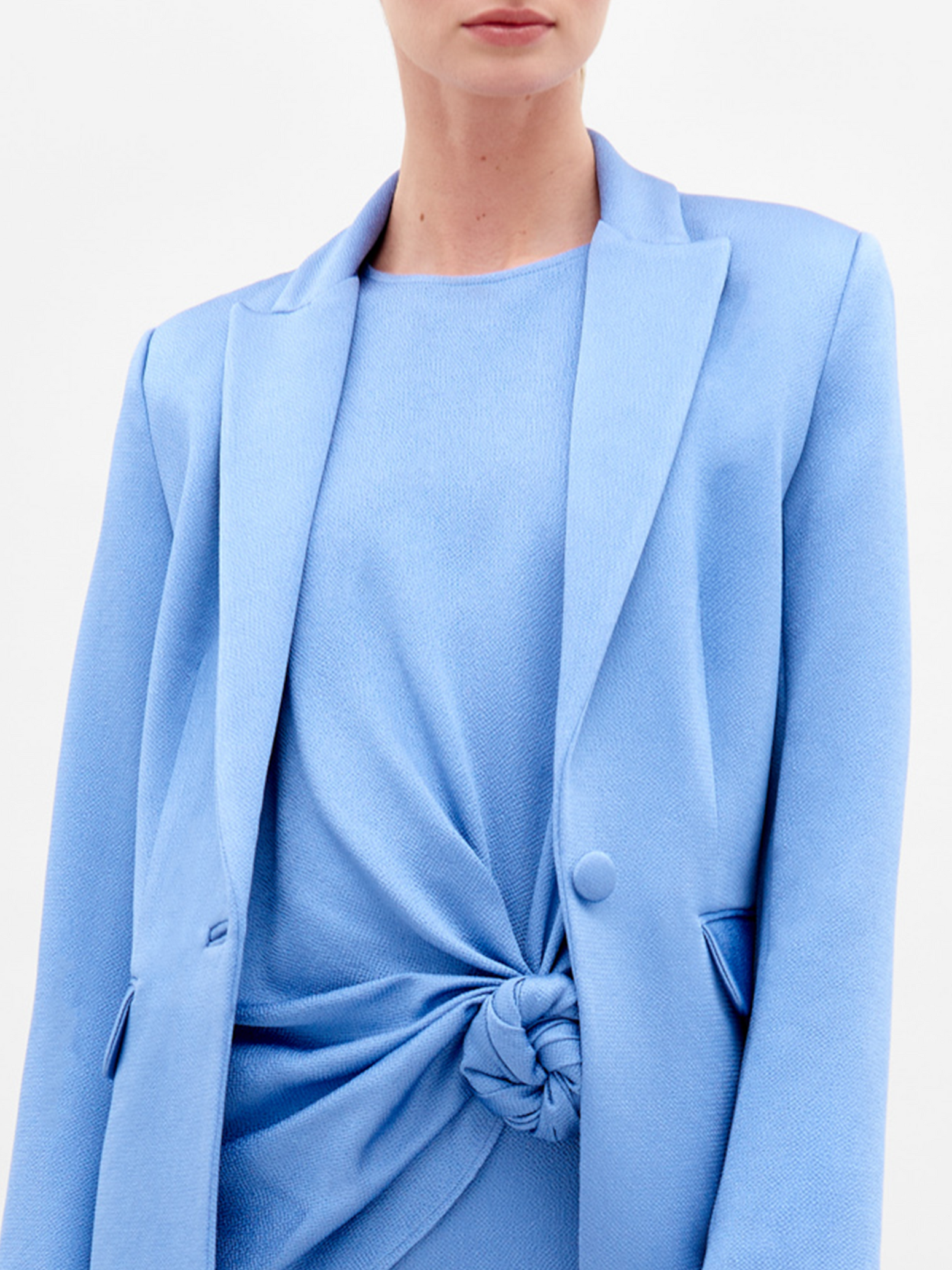 Wearing the elegant Rebeca Jacket by SVT DESIGNS, a light blue hammered satin blazer with a single-button front over a matching dress with waist knot detail. The plain white background highlights the Italian textile, while the person's face is partially cropped in the image.