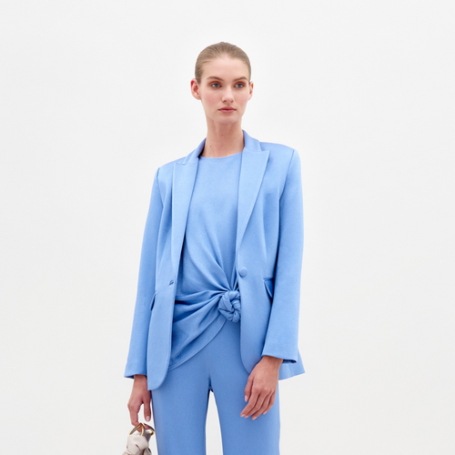 Wearing the Rebeca Jacket from SVT DESIGNS, a person dons a light blue pantsuit with a knotted design at the front, paired with matching luxurious Italian textile pants. They carry a small, light handbag in their left hand against a plain white background.