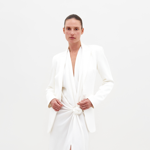 A person dressed in SVT DESIGNS' Rebeca Jacket, a white hammered satin blazer with notched lapels, over a matching dress featuring a knotted waist, stands against a plain white background, gazing directly at the camera with a neutral expression.