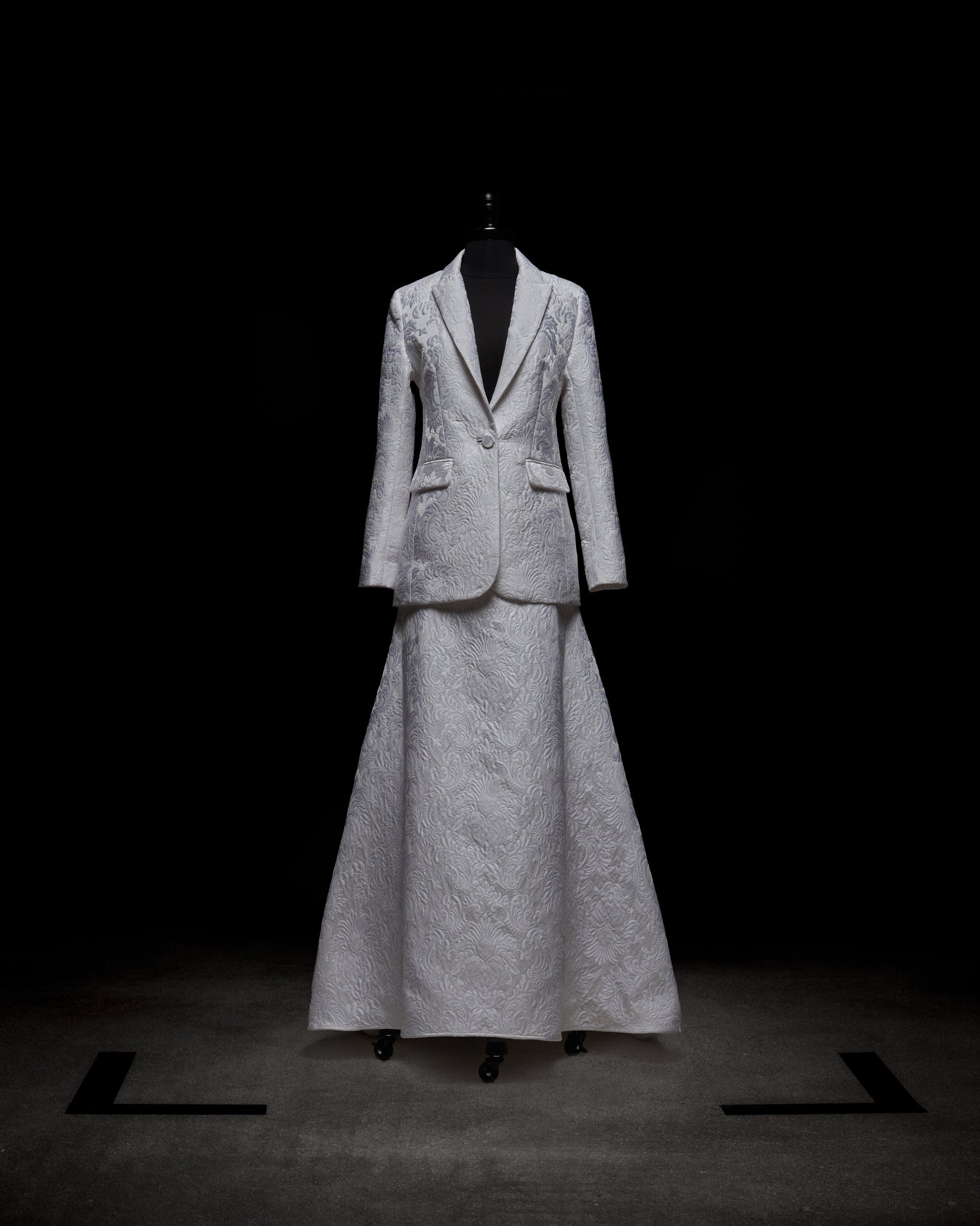 A mannequin features a chic ensemble from SVT DESIGNS, showcasing the 'Imola Skirt,' a long, textured white piece paired with an elegantly embroidered matching blazer against a dark backdrop.