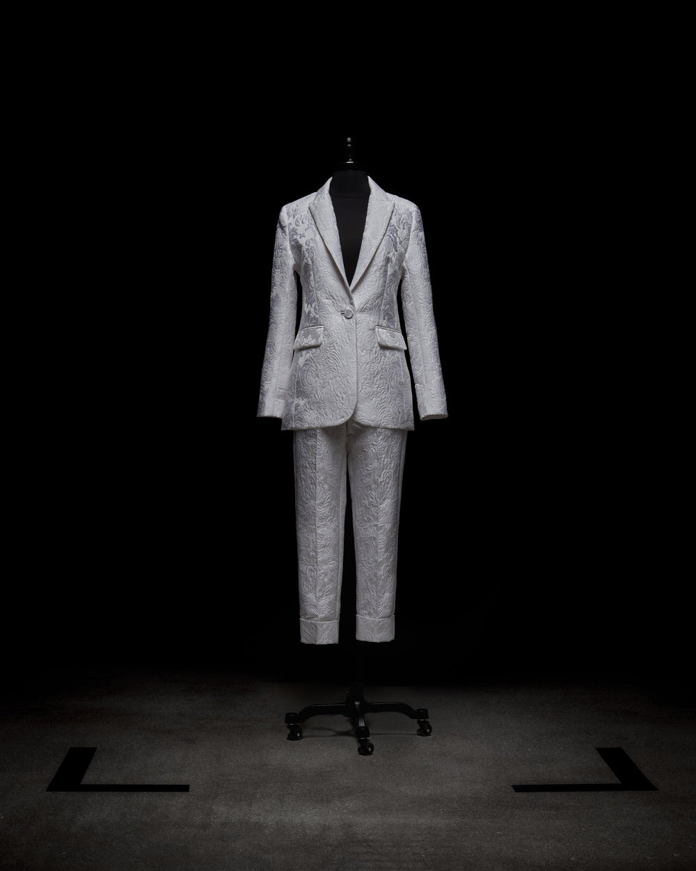 A silver patterned Rebeca Jacket by SVT DESIGNS is displayed on a mannequin against a black background, featuring a single-breasted style with notched lapels and matching trousers. The lighting accentuates the fabric's texture and intricate design.