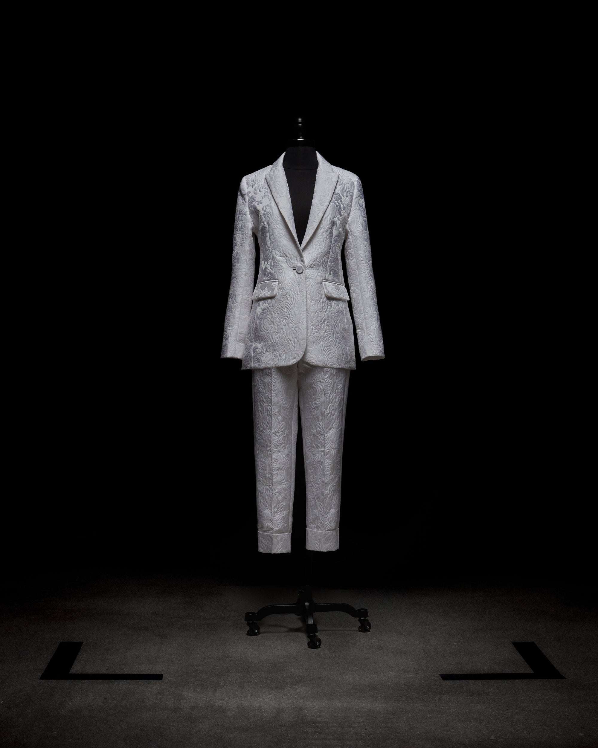 A silver patterned Rebeca Jacket by SVT DESIGNS is displayed on a mannequin against a black background, featuring a single-breasted style with notched lapels and matching trousers. The lighting accentuates the fabric's texture and intricate design.