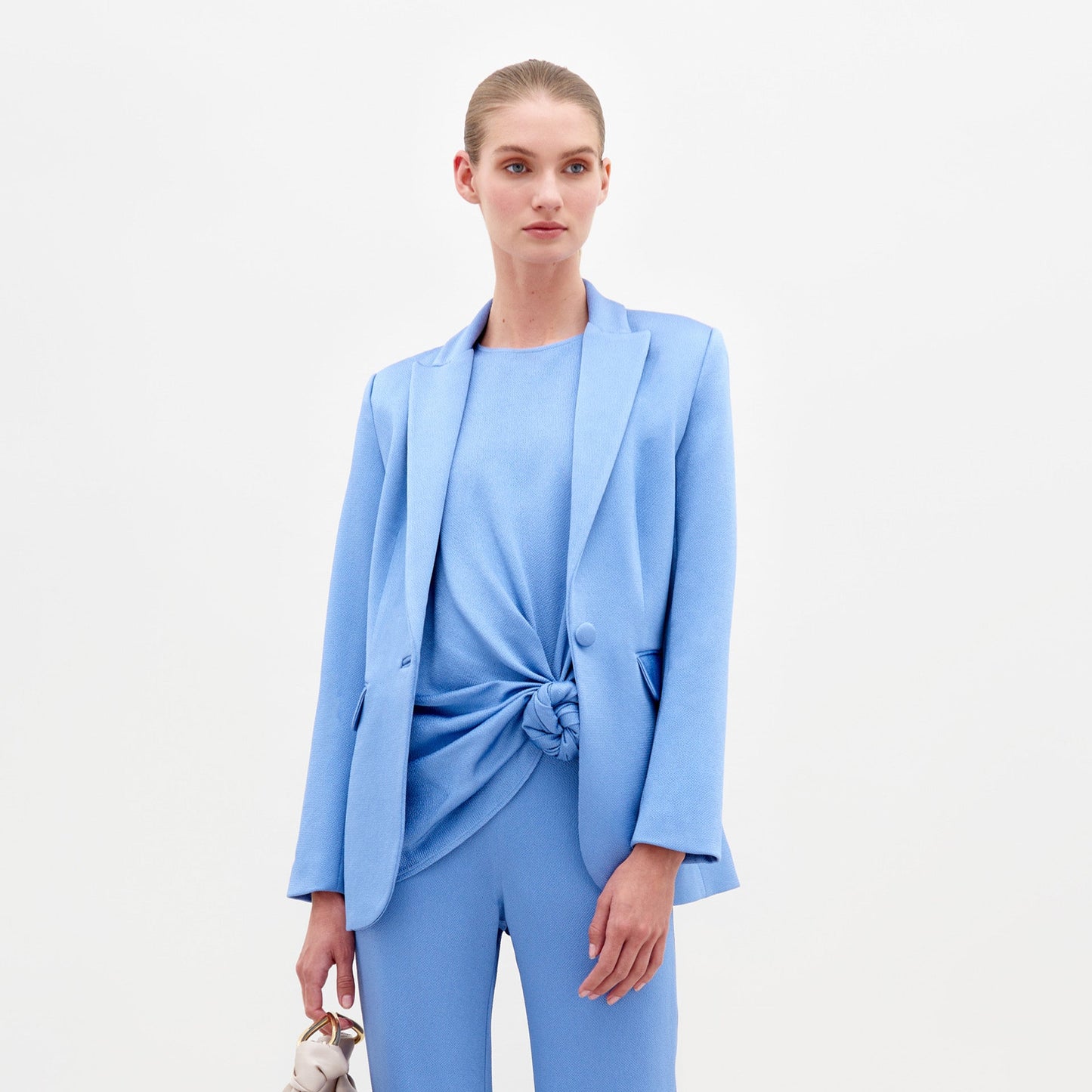 A woman in a light blue Rebeca Jacket with hammered satin, single-button closure, and matching knotted top and pants holds sunglasses against a plain white background.