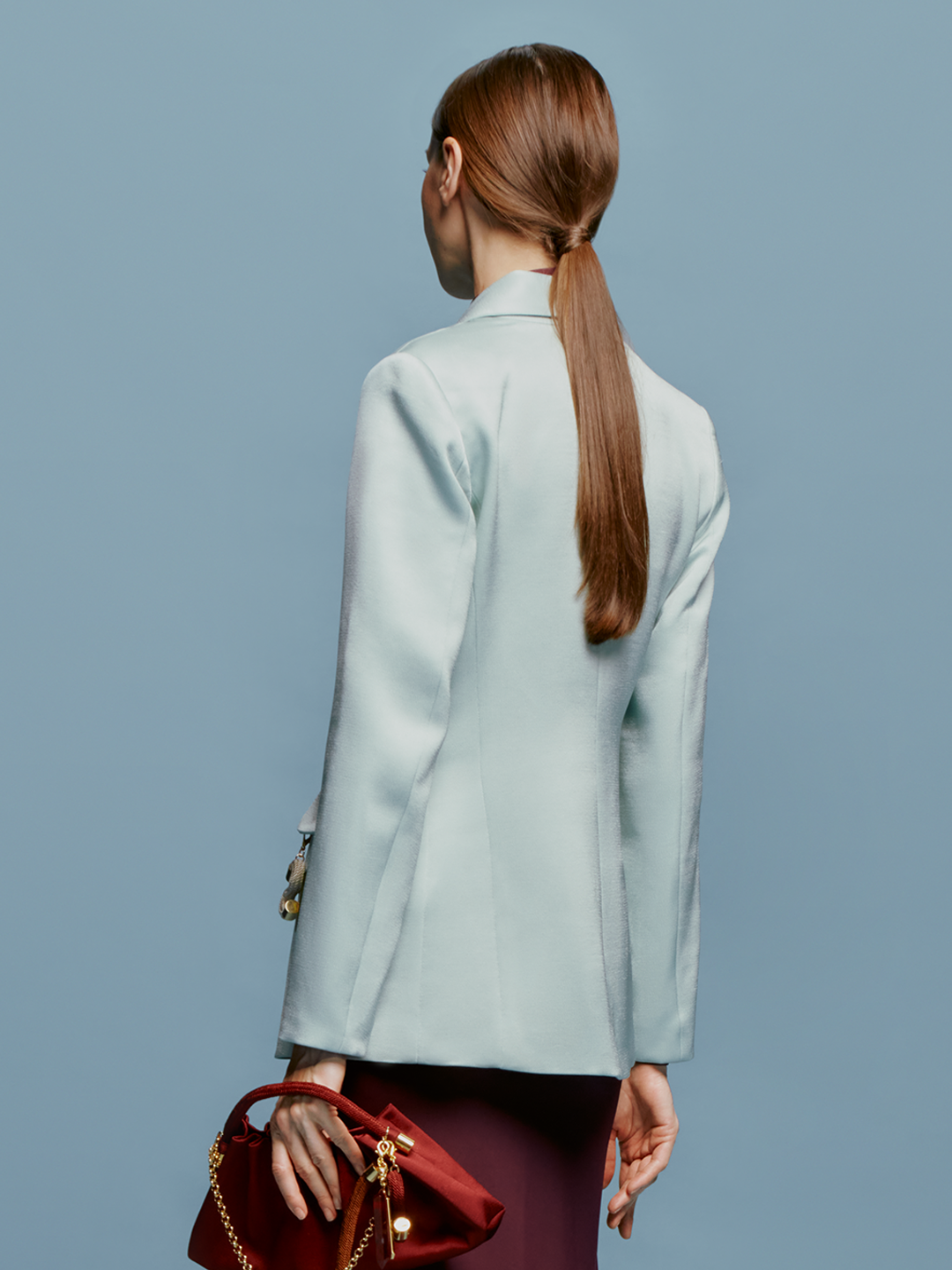 A woman in a turquoise Rebeca Jacket over a burgundy top stands against a light blue background. She has straight, pulled-back hair and an "S" charm with an attached trinket hanging from her jacket pocket, ready to make a statement on September 2nd.