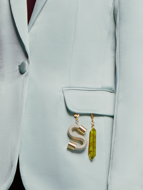 A woman in a turquoise Rebeca Jacket over a burgundy top stands against a light blue background. She has straight, pulled-back hair and an "S" charm with an attached trinket hanging from her jacket pocket, ready to make a statement on September 2nd.