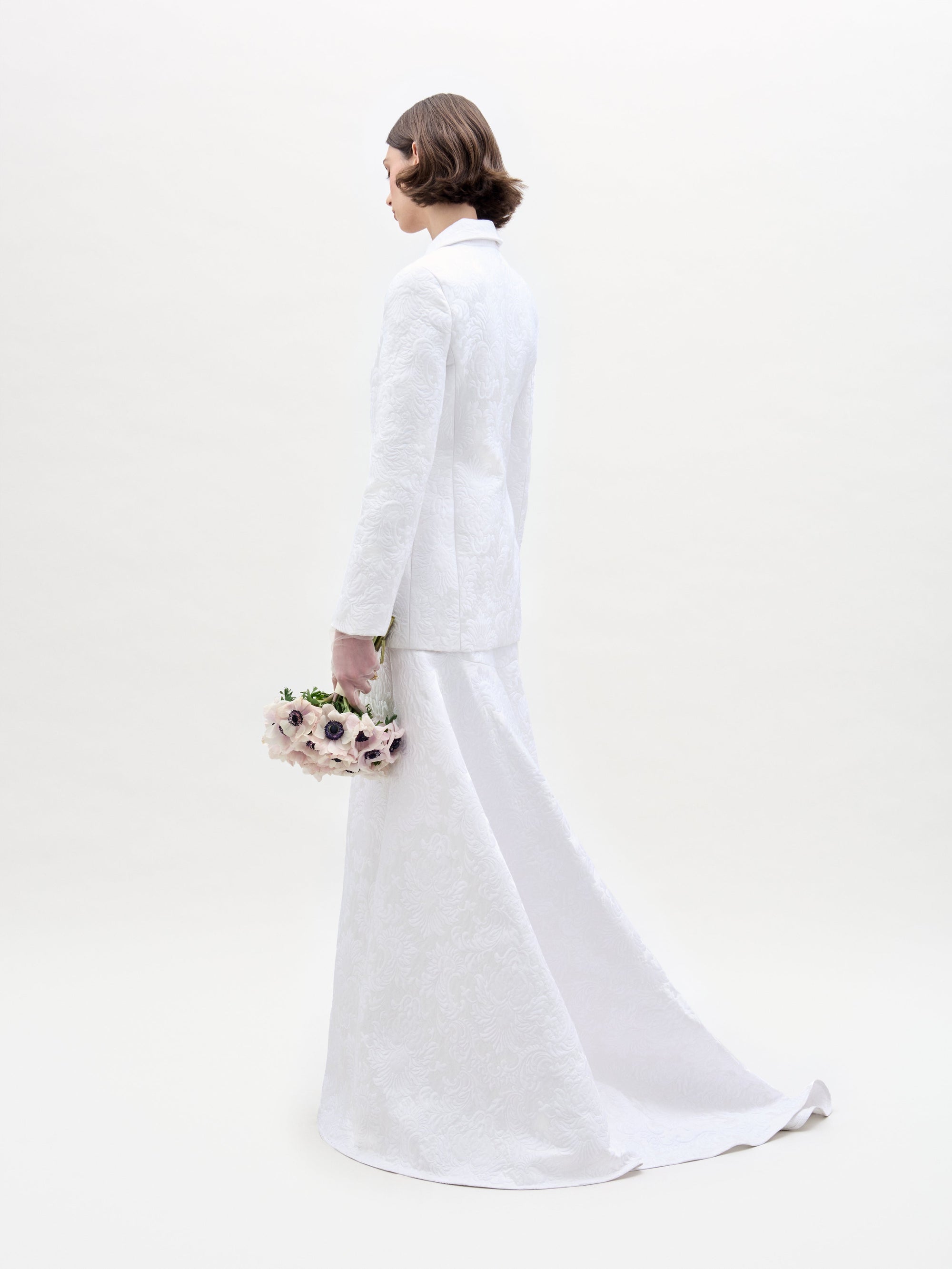 A person with short brown hair faces away from the camera, dressed in a white textured suit featuring the Imola Skirt by SVT DESIGNS. They hold a bouquet of light-colored flowers with dark centers against a plain white backdrop.