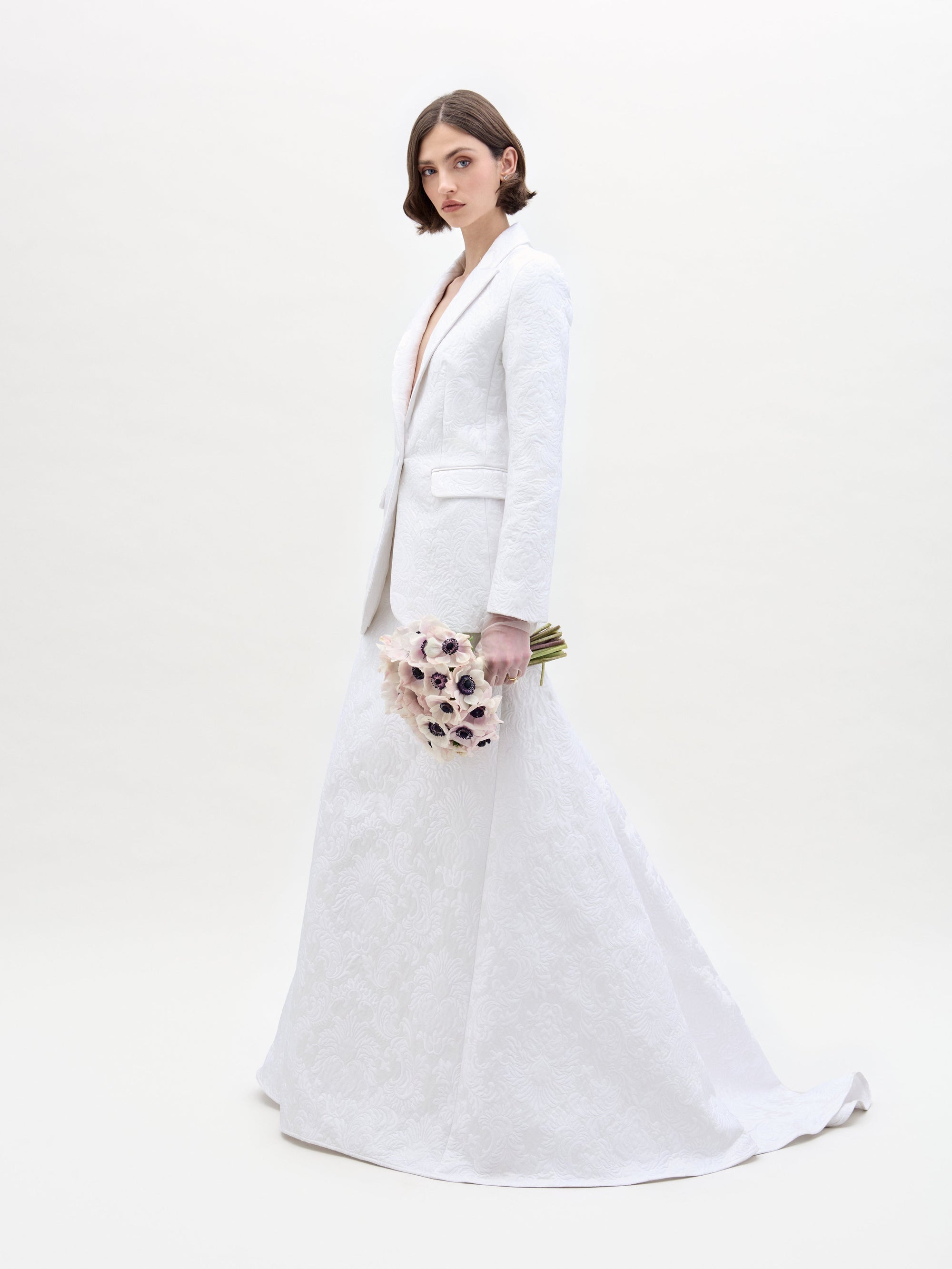 Wearing the Imola Skirt by SVT DESIGNS, an individual in a textured white suit dress stands before a plain backdrop, holding a bouquet of light pink flowers with dark centers. The attire features a blazer top and long skirt.