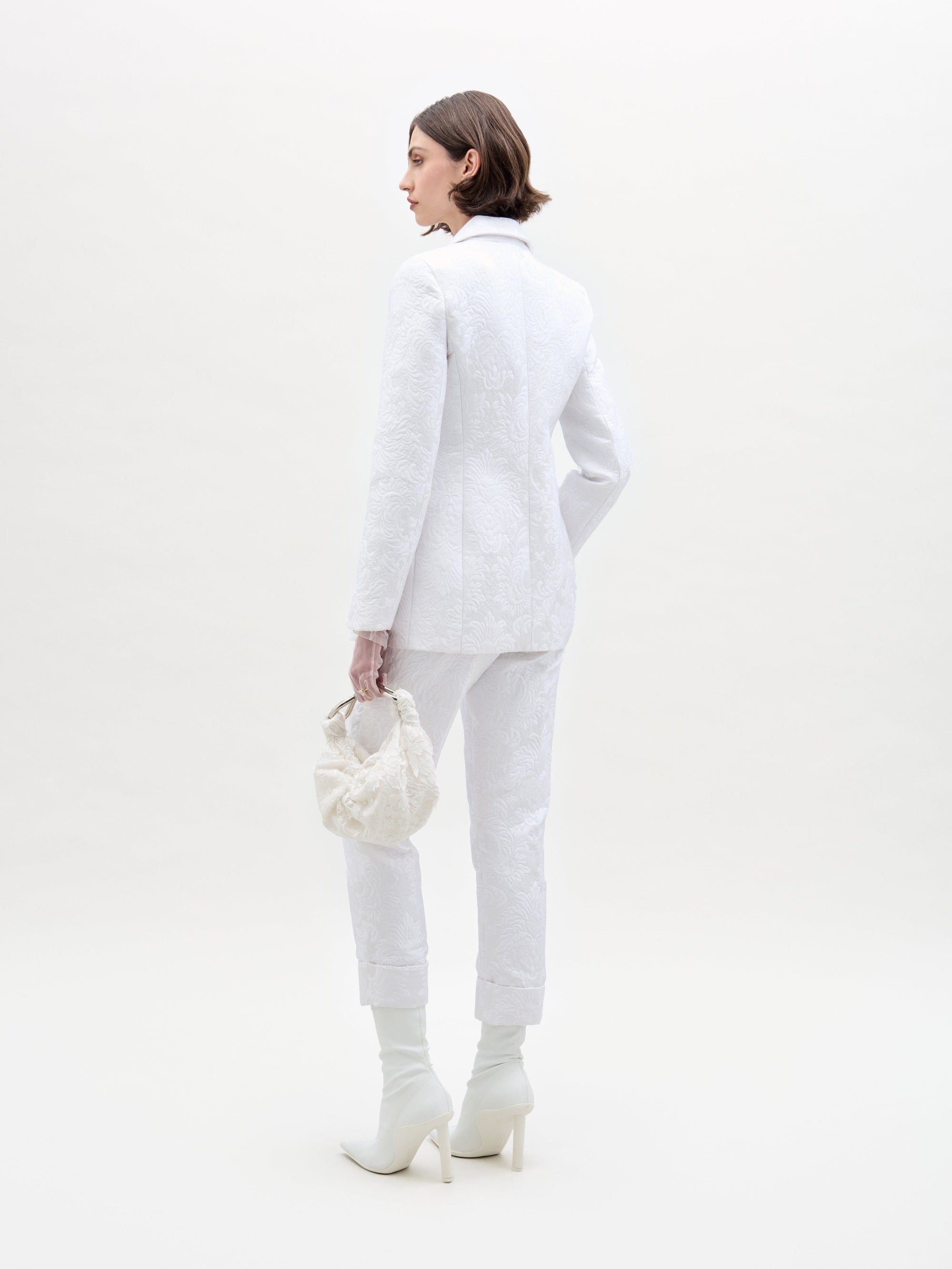 A woman with short hair faces away, dressed in a textured all-white ensemble consisting of the Rebeca Jacket and cropped pants by SVT DESIGNS. She carries a small white handbag, wears white heeled boots, and stands against a plain light background.