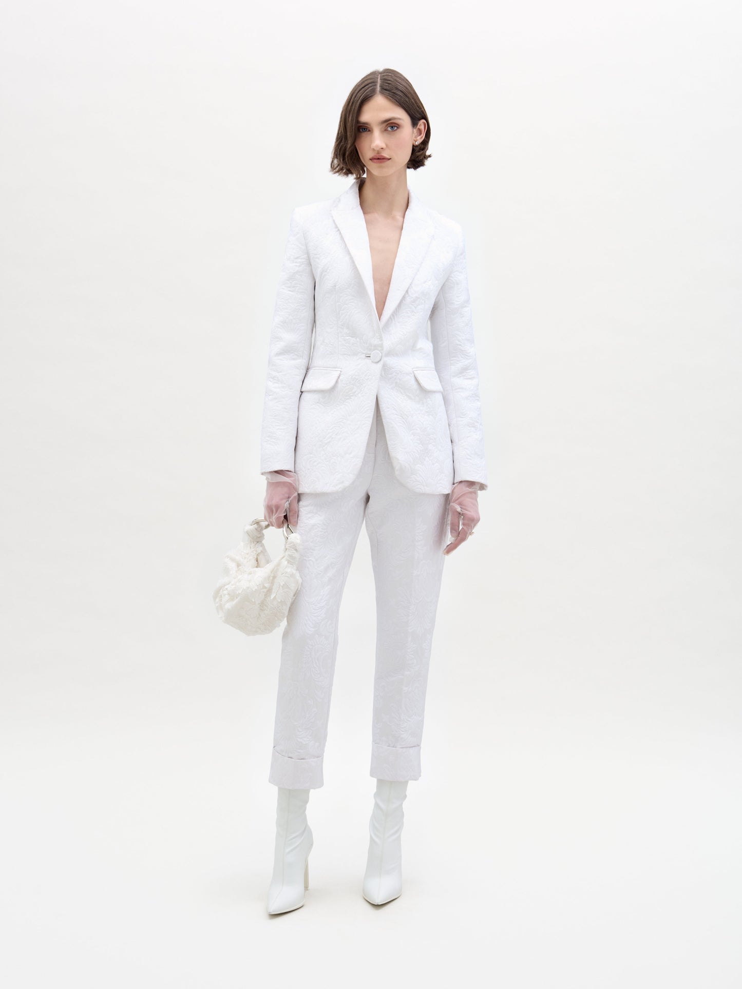 A person stands against a plain background, wearing SVT DESIGNS' textured Moad Pant and matching deep neckline suit, styled with a small white handbag and white ankle boots. Their hair is in a sleek bob.