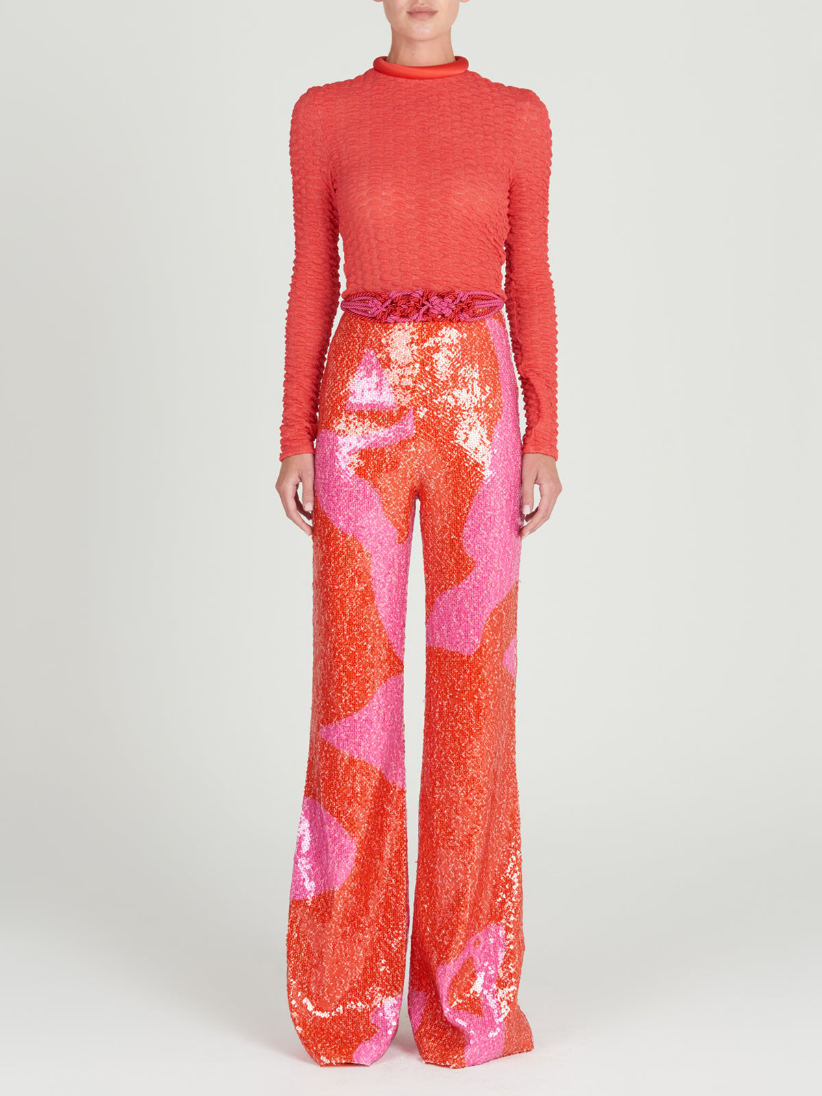 A person stands facing forward in the Jari Top, a red long-sleeve embossed jacquard piece, paired with high-waisted, wide-leg sequin pants featuring orange and pink abstract patterns, against a plain light background.