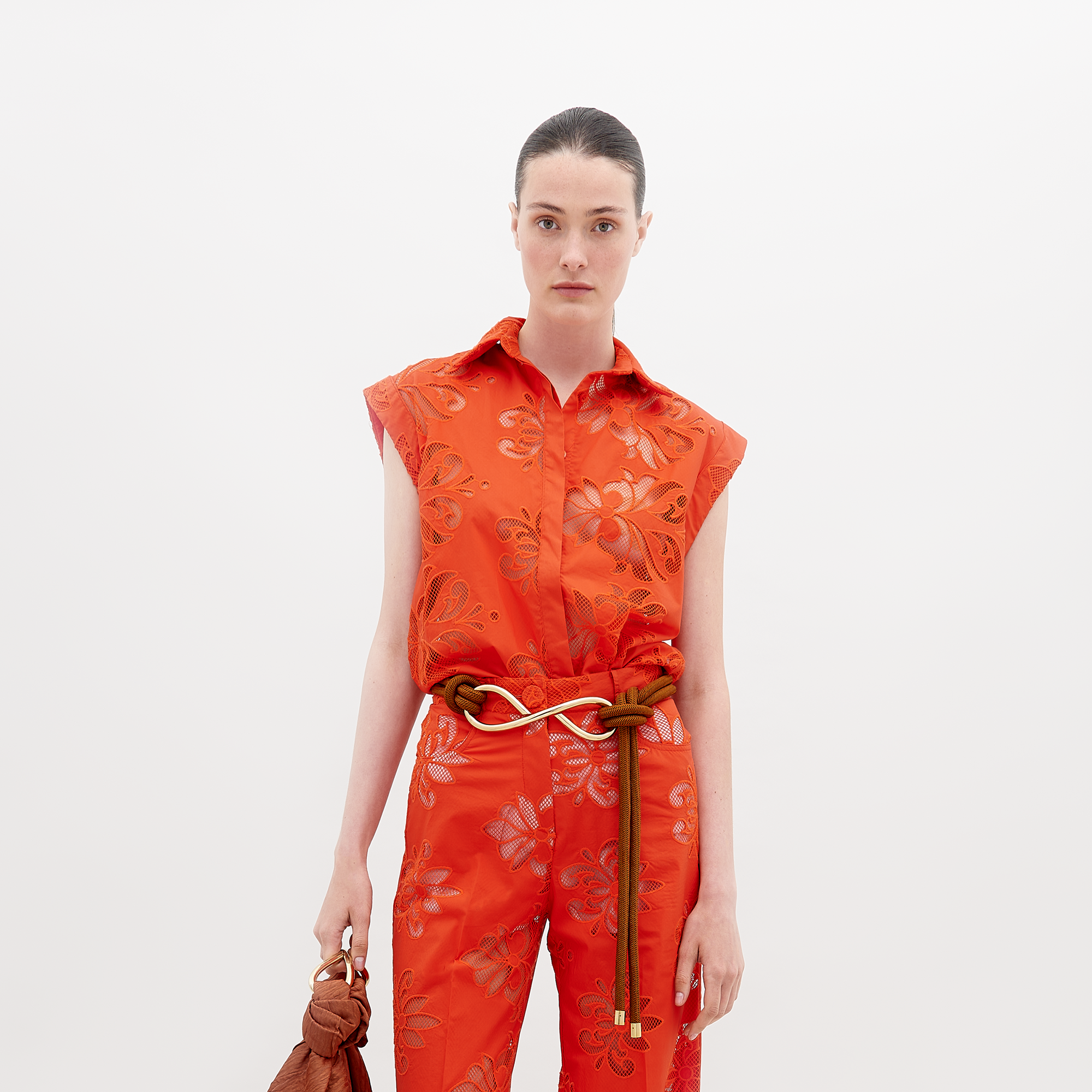 A person with dark hair is in a bright orange floral-patterned jumpsuit. They wear a brown braided belt and hold an orange bag against a plain white backdrop. Featured items include the Rina Blouse by SVT DESIGNS.