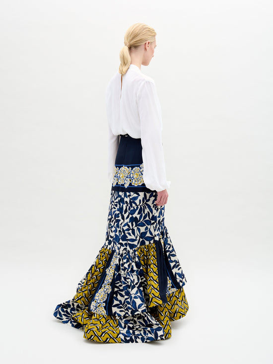 A blonde-haired person in a ponytail wears the Elliot Skirt by SVT DESIGNS, a long maxi with blue, yellow, and white patterns made from Italian fabric, paired with a white blouse. They stand against a plain, light background, facing slightly away from the camera.