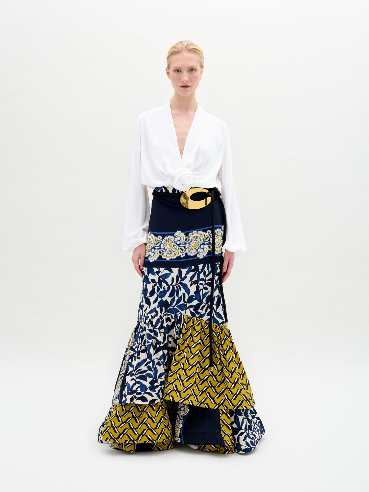 A person poses against a plain backdrop, wearing the Elliot Skirt from SVT DESIGNS. This vibrant maxi skirt features blue and yellow patterns on exquisite Italian fabric, with geometric and floral designs. It includes a large belt with a gold buckle and is paired with a white long-sleeve blouse.