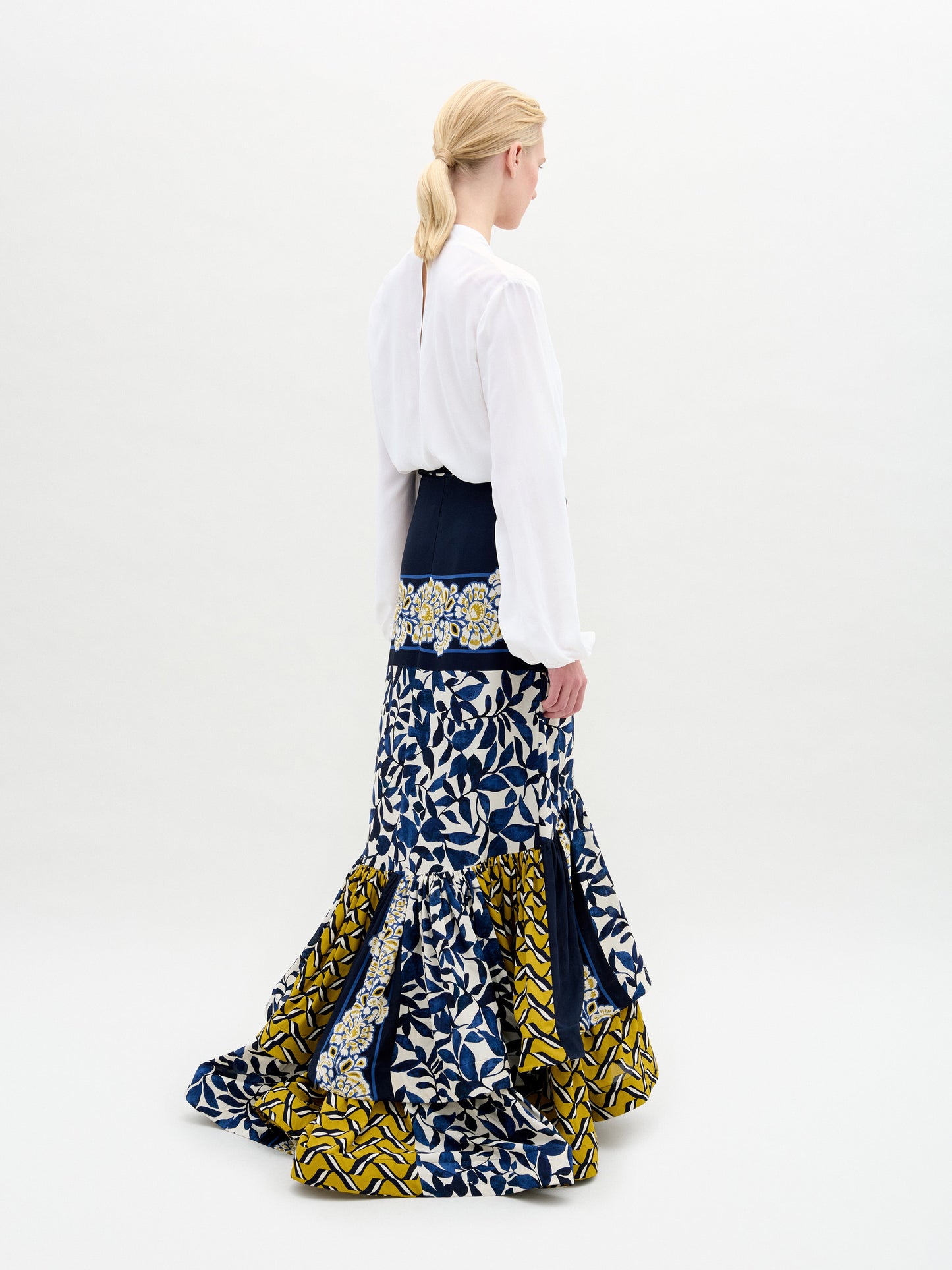 A woman with blonde hair in a low ponytail stands facing away, wearing the Rocio Blouse by SVT DESIGNS tucked into a high-waisted skirt made from Italian fabric with blue, white, and yellow geometric and floral patterns.