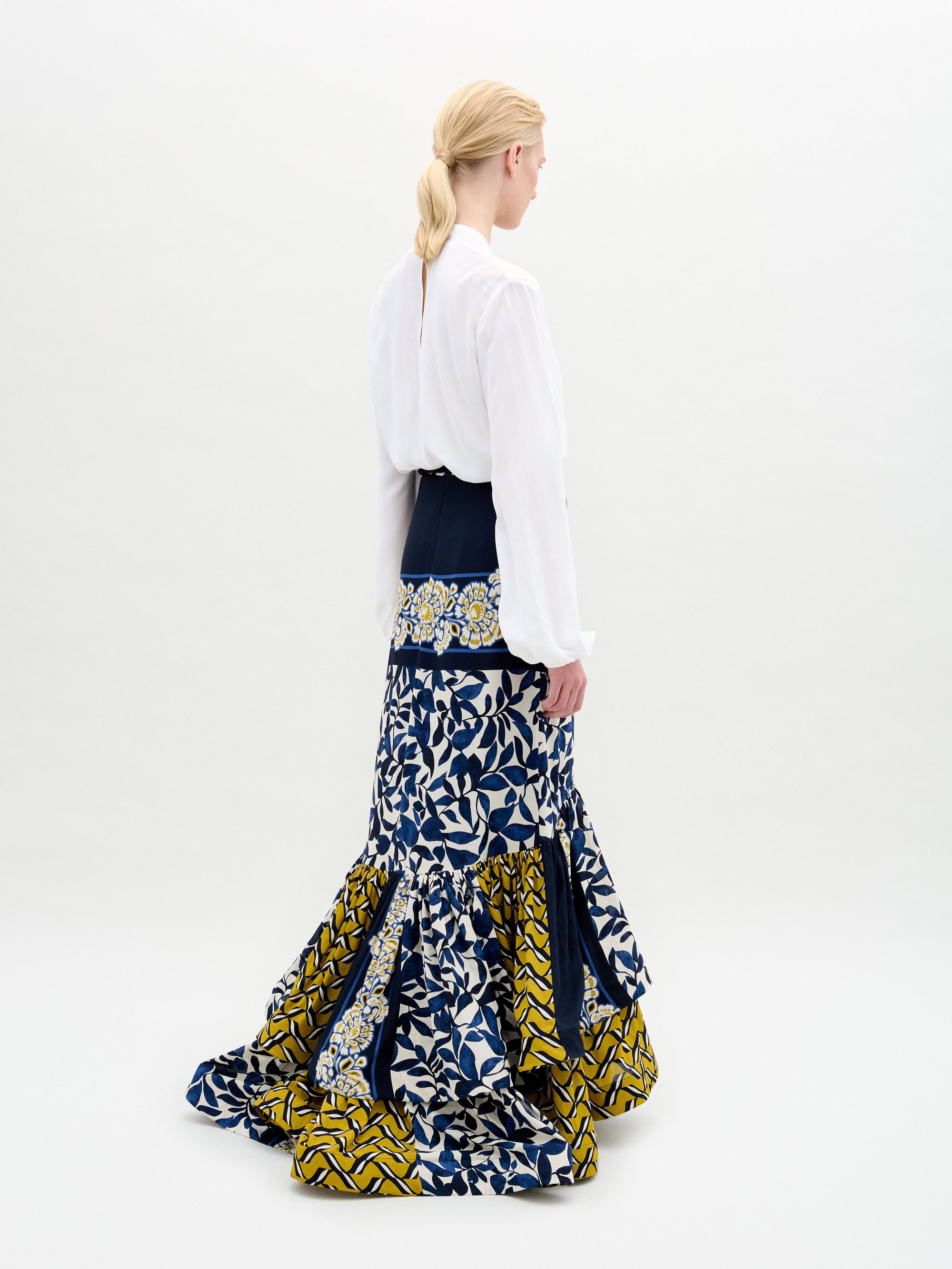 A woman with blonde hair in a low ponytail stands facing away, wearing the Rocio Blouse by SVT DESIGNS tucked into a high-waisted skirt made from Italian fabric with blue, white, and yellow geometric and floral patterns.