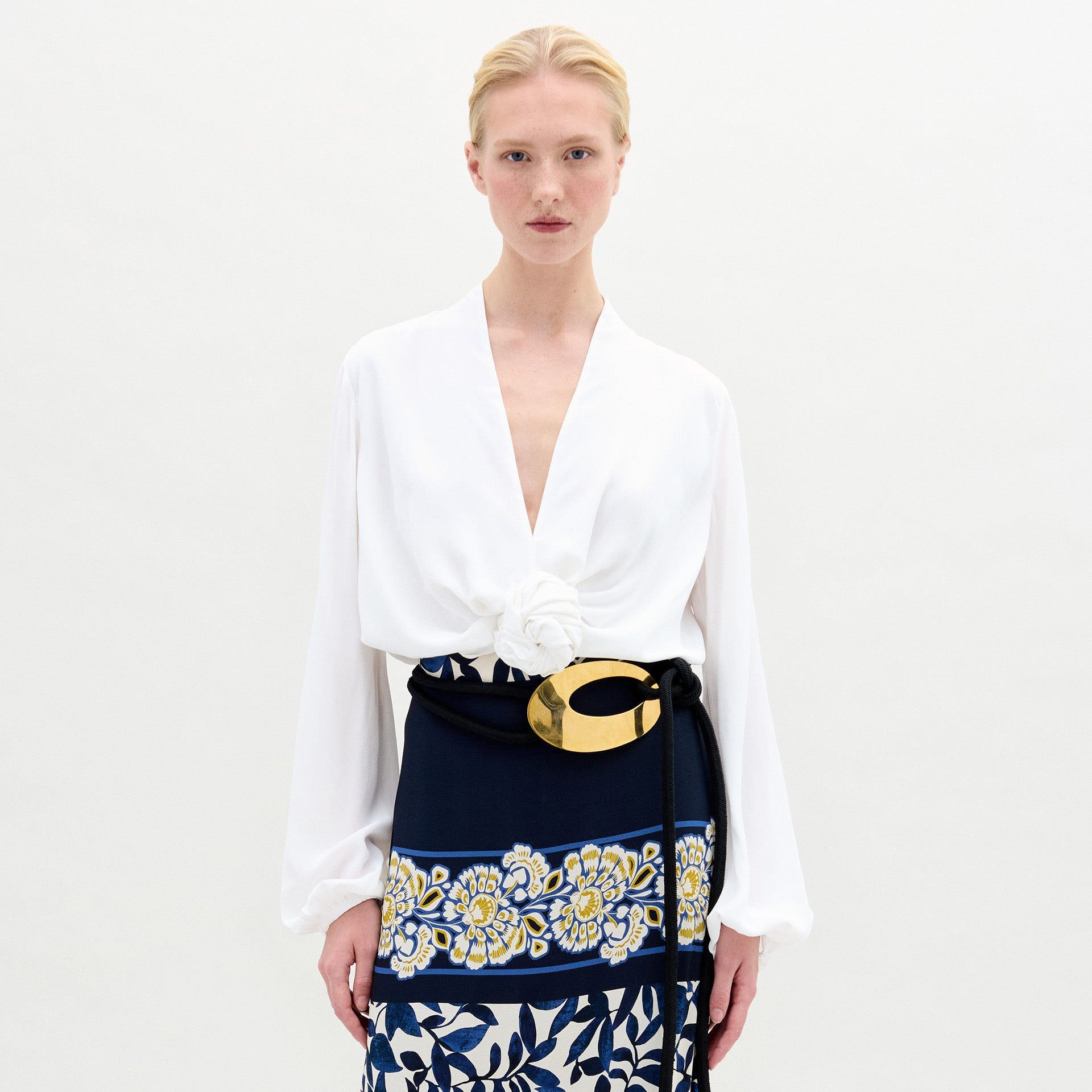 A person with blonde hair wears the SVT DESIGNS Rocio Blouse—white, long-sleeved, with a deep V-neck and flower detail at the waist—paired with a navy skirt featuring yellow and white florals and an asymmetrical hem. The backdrop is plain and light.