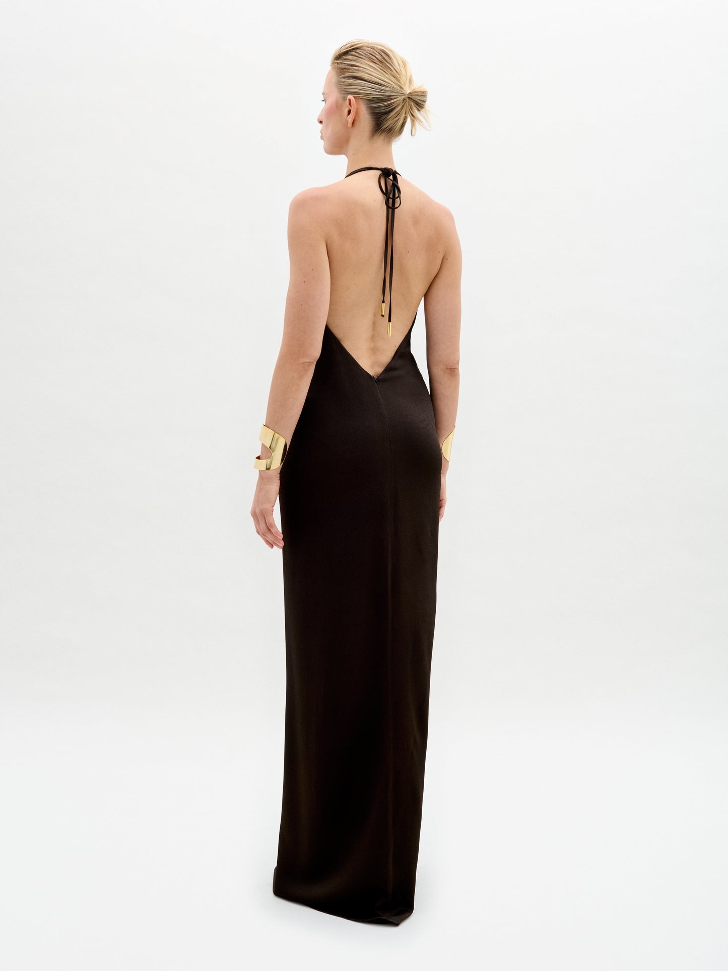 A woman with short blonde hair wears the SVT DESIGNS Rowan Dress, a sleeveless hammered satin maxi dress elegantly tied at the halter neckline. Gold bracelets adorn her wrists as she stands against a plain white background, facing away from the camera.