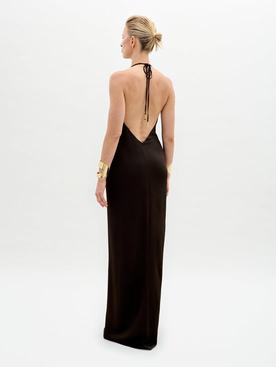 A woman with short blonde hair wears the SVT DESIGNS Rowan Dress, a sleeveless hammered satin maxi dress elegantly tied at the halter neckline. Gold bracelets adorn her wrists as she stands against a plain white background, facing away from the camera.