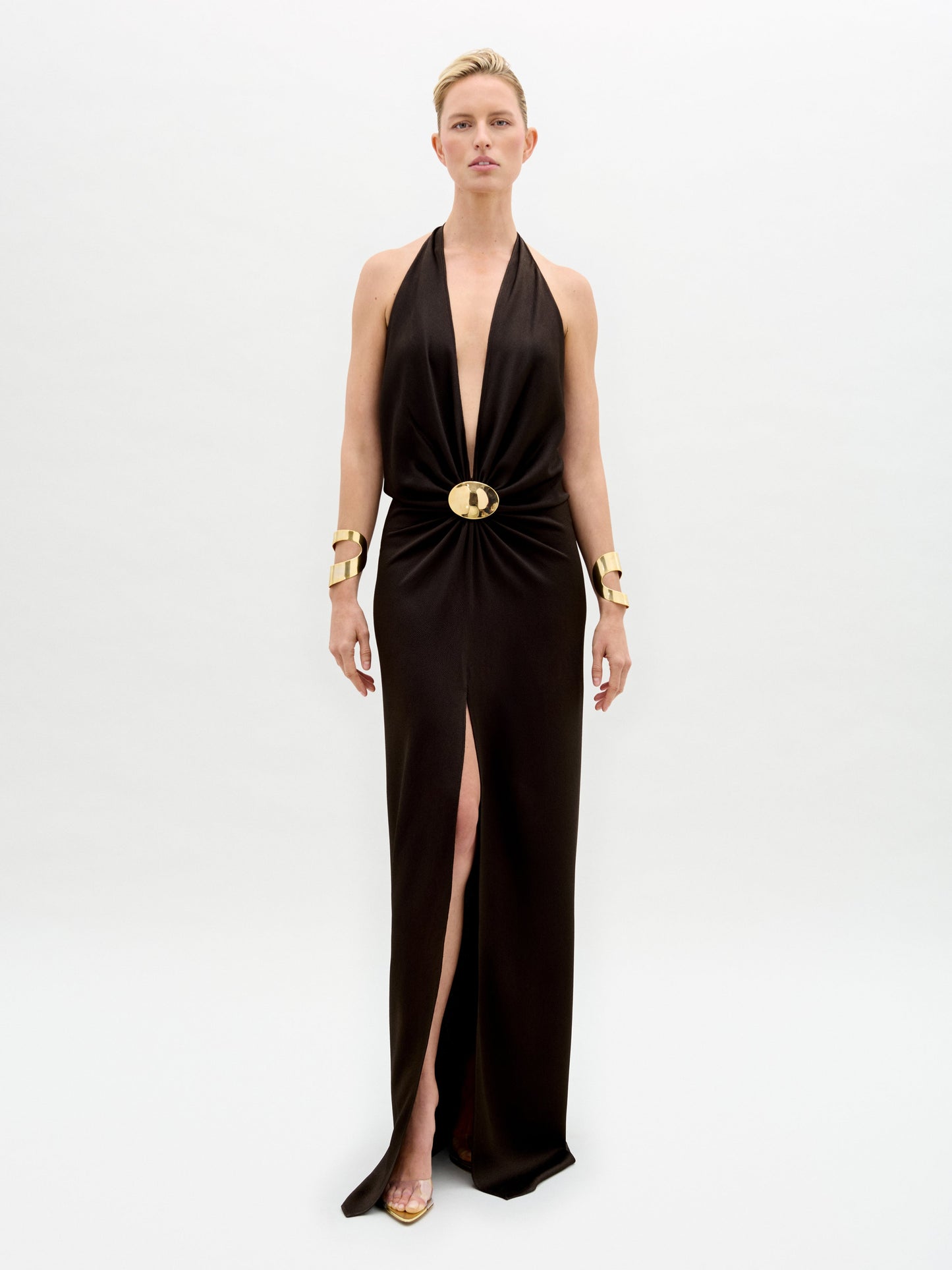 A person stands against a white background wearing the Rowan Dress by SVT DESIGNS. This black hammered satin maxi dress features a deep halter neckline, high slit, and circular waist embellishment. They accessorize with gold bracelets and heels.