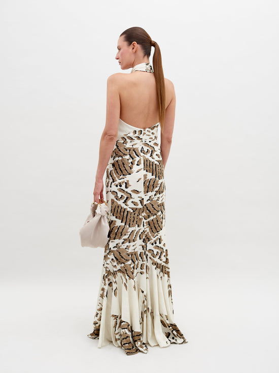 A woman stands in a Raisa Dress Beige Abstract Animal Print with a halter neckline, holding a small, light-colored handbag. She has a neutral expression and stands against a plain white background. This chic ensemble is available for pre-order, with a ship by date of November 15th, 2024.