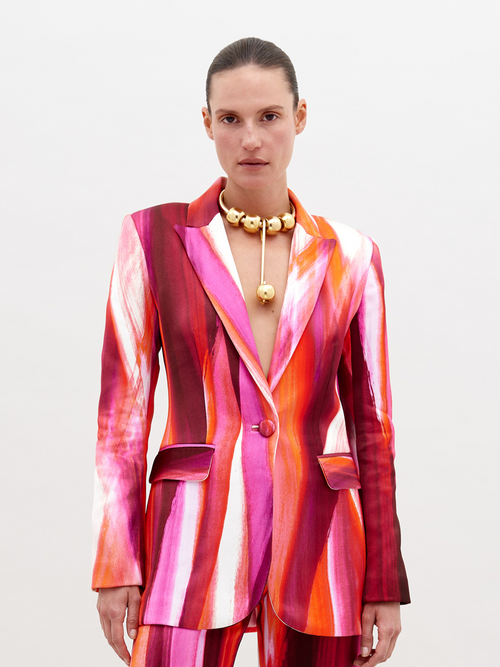 Woman wearing a vibrant, multi-colored striped Rebeca Jacket Magenta Abstract Waves with a deep neckline, accessorized with a chunky gold choker necklace, standing against a plain white background. Pre-order now and have it ship by date of November 15th, 2024.