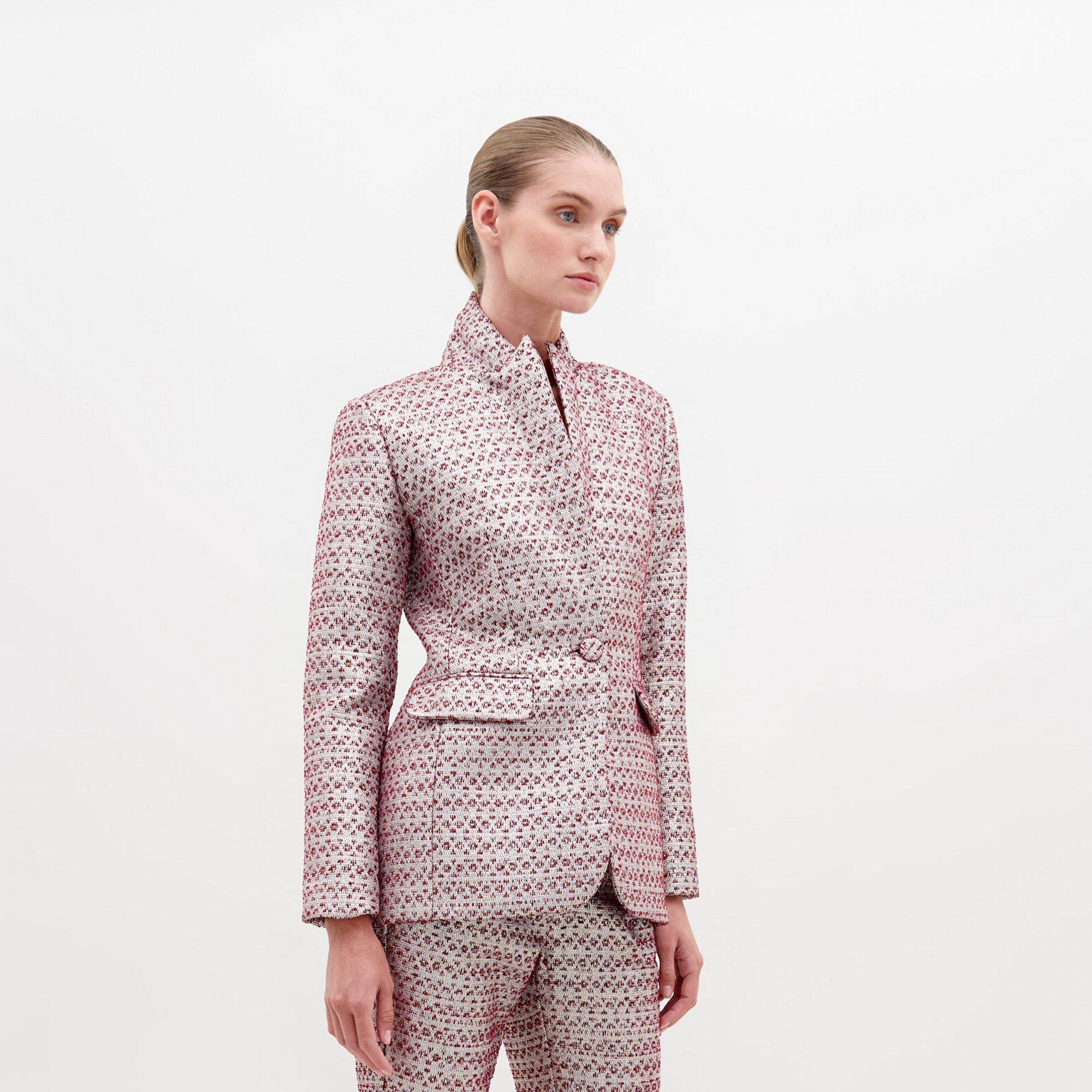 A person stands against a plain white background, wearing the SVT DESIGNS Rebeca Jacket Metallic Burgundy. This structured fit jacket is crafted from Italian textile with a tailored silhouette, high collar, belted waist, and red and white geometric pattern.